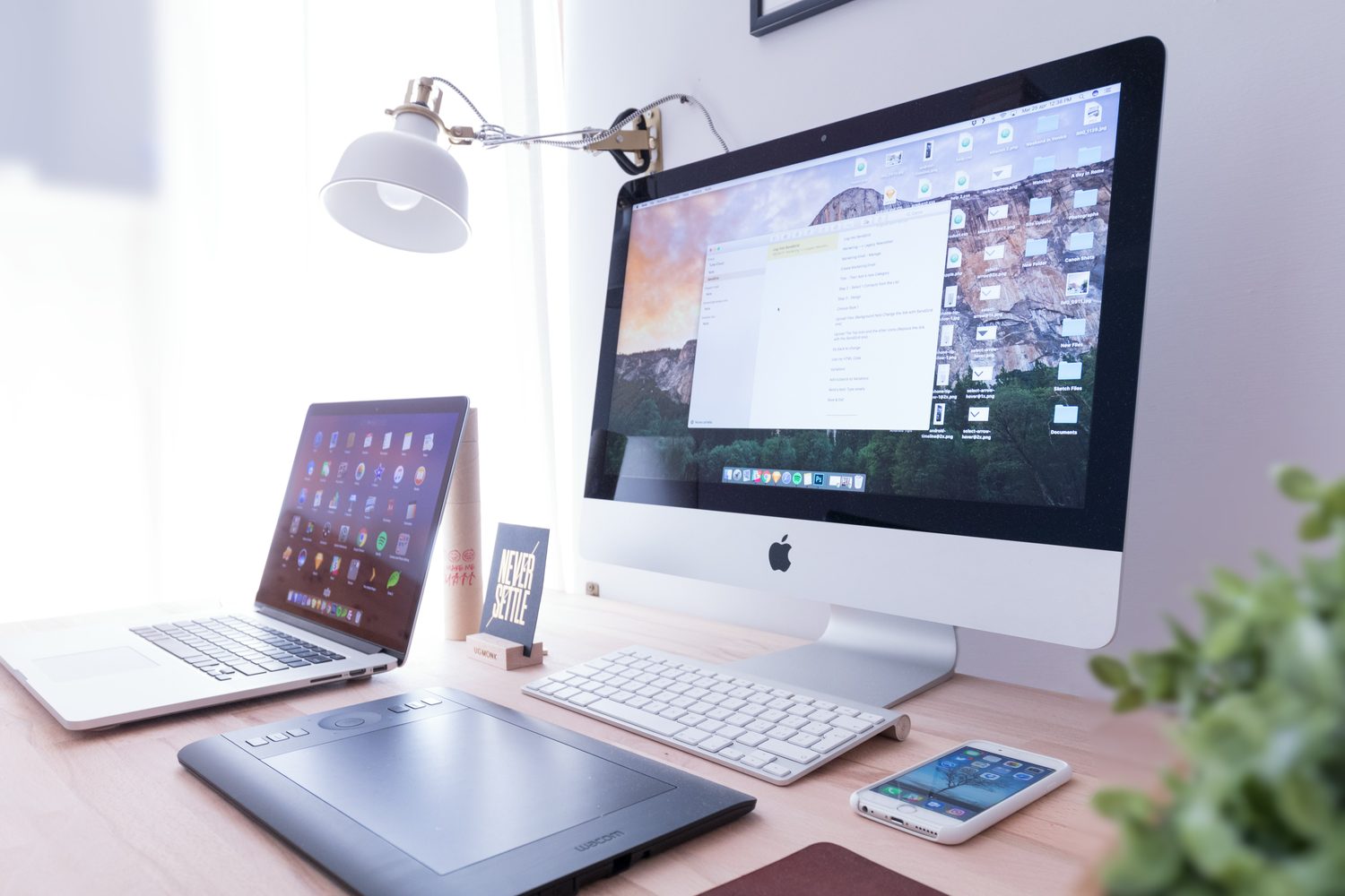 A tidy desk with a large monitor, a laptop, a graphics tablet and a phone arranged under an angled lamp