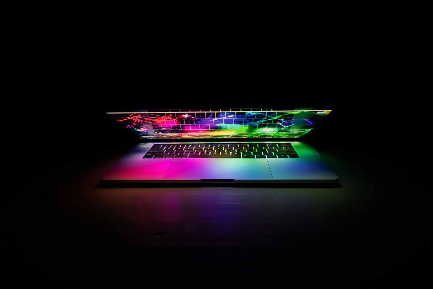 A laptop open in a dark room with saturated colour spilling from the screen across the keyboard